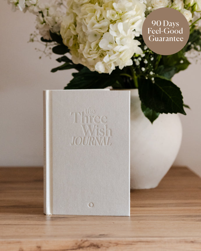 White premium journal titled "The Three Wish Journal" on a wooden table, next to a vase of white flowers - for calm, connection and emotional resilience through kindness.